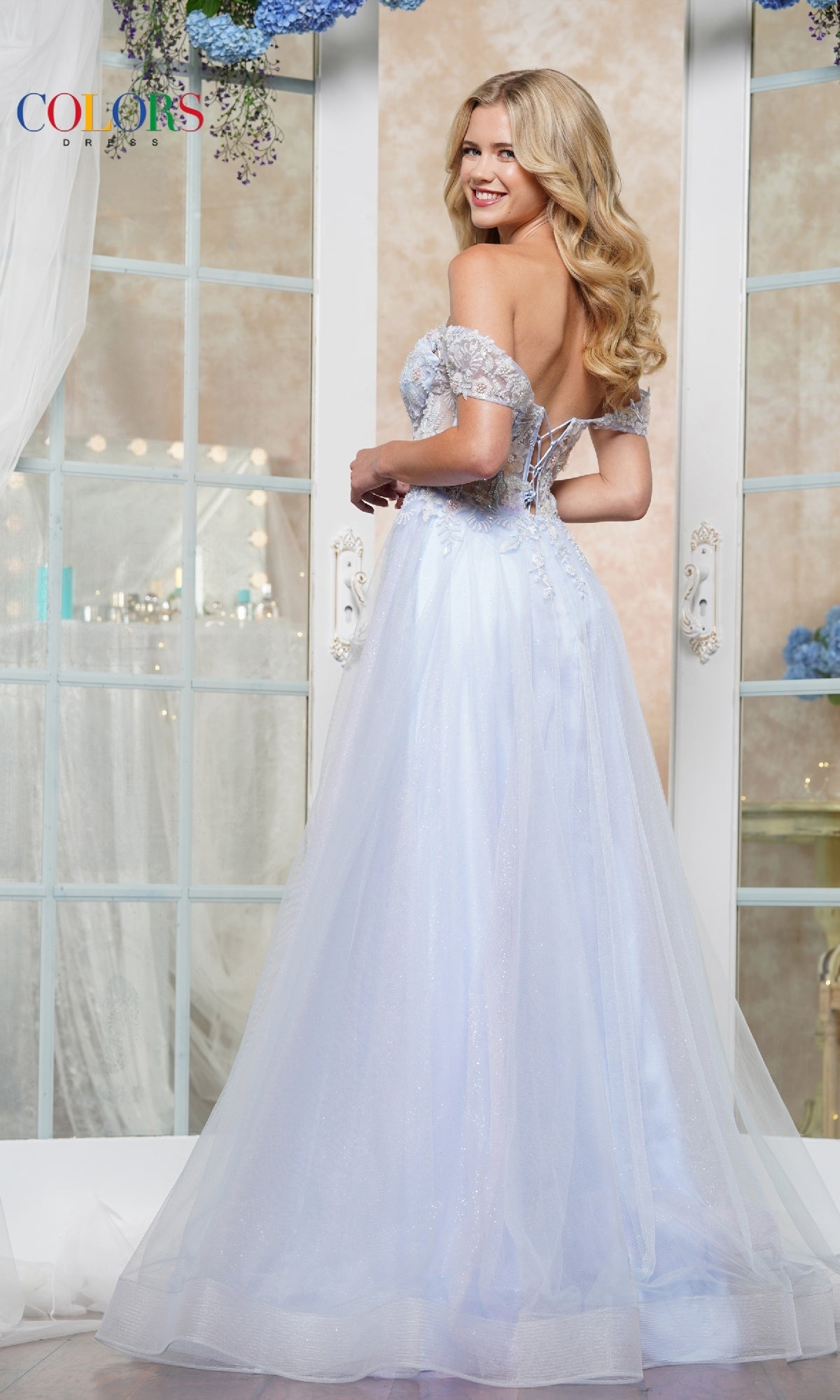 A woman smiles over her shoulder in front of large windows, wearing the Off-Shoulder Glitter Tulle Prom Ball Gown: Colors Dress 3498 with a flowing pale blue skirt.